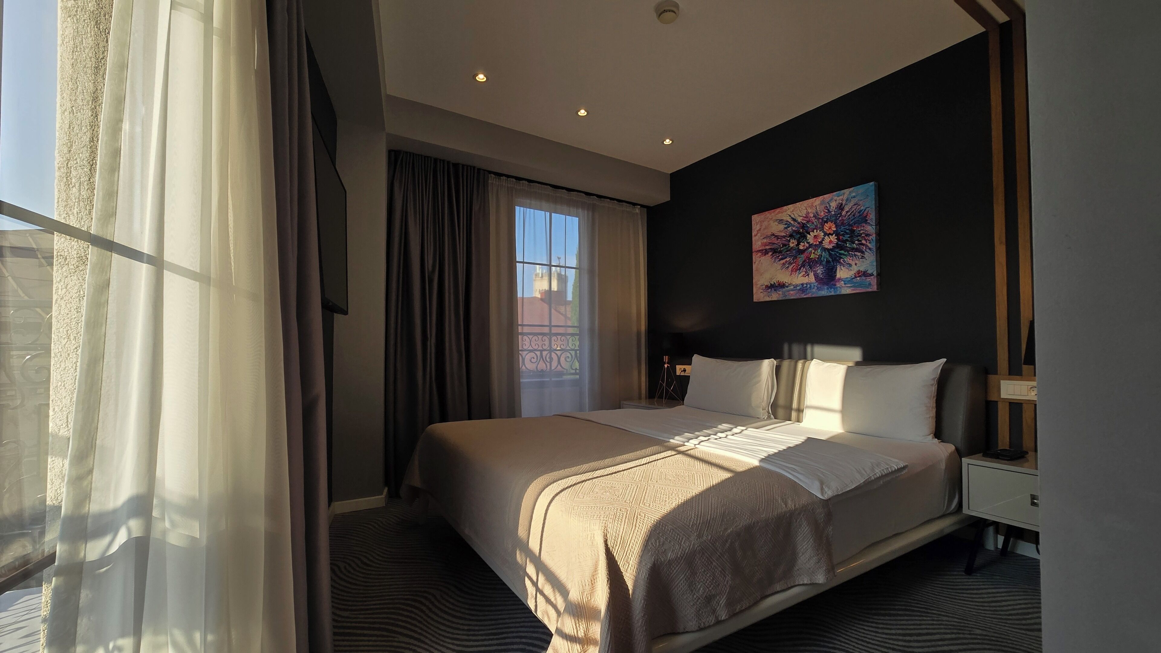 A hotel room with a large bed, a nightstand, a painting on the wall, and a view of a building through the window.