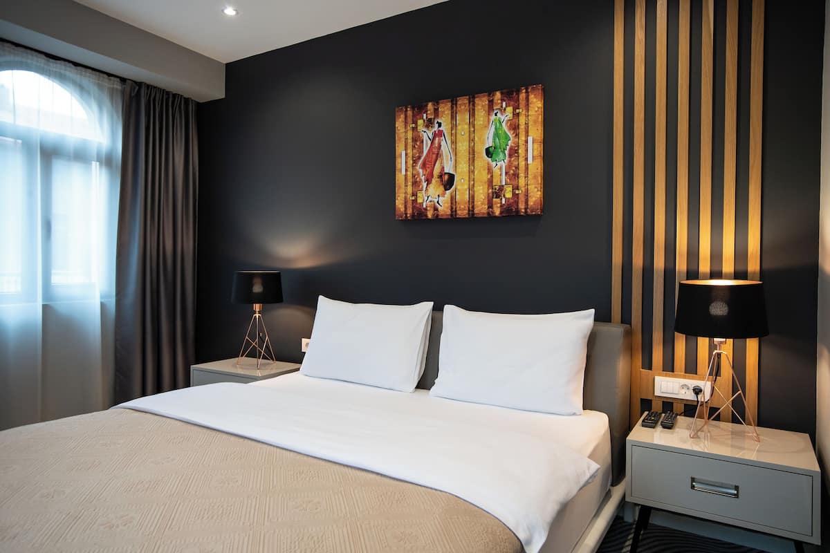 A modern hotel room with a large bed, two bedside tables with lamps, a painting on the wall, and a window with curtains.