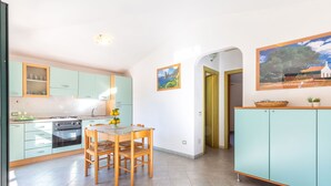 Cuina amb taula i caires, fogó i armaris. Hi ha un quadre a la paret i una porta que condueix a una altra habitació.