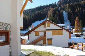 A chalet-style building with a wooden staircase leading to a balcony, surrounded by snow-covered ground and forested mountains in the background. - Casa Mazzoni (Val di Zoldo)