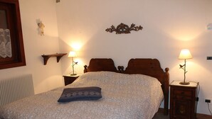 A bedroom with a large bed, two bedside tables with lamps, a wooden headboard, a wall-mounted shelf, and a radiator. - Casa Mazzoni (Val di Zoldo)