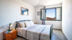 A bedroom with a bed, nightstand, lamp, and a window with a view of buildings. - Residence Verde Navarrese B (Baunei)