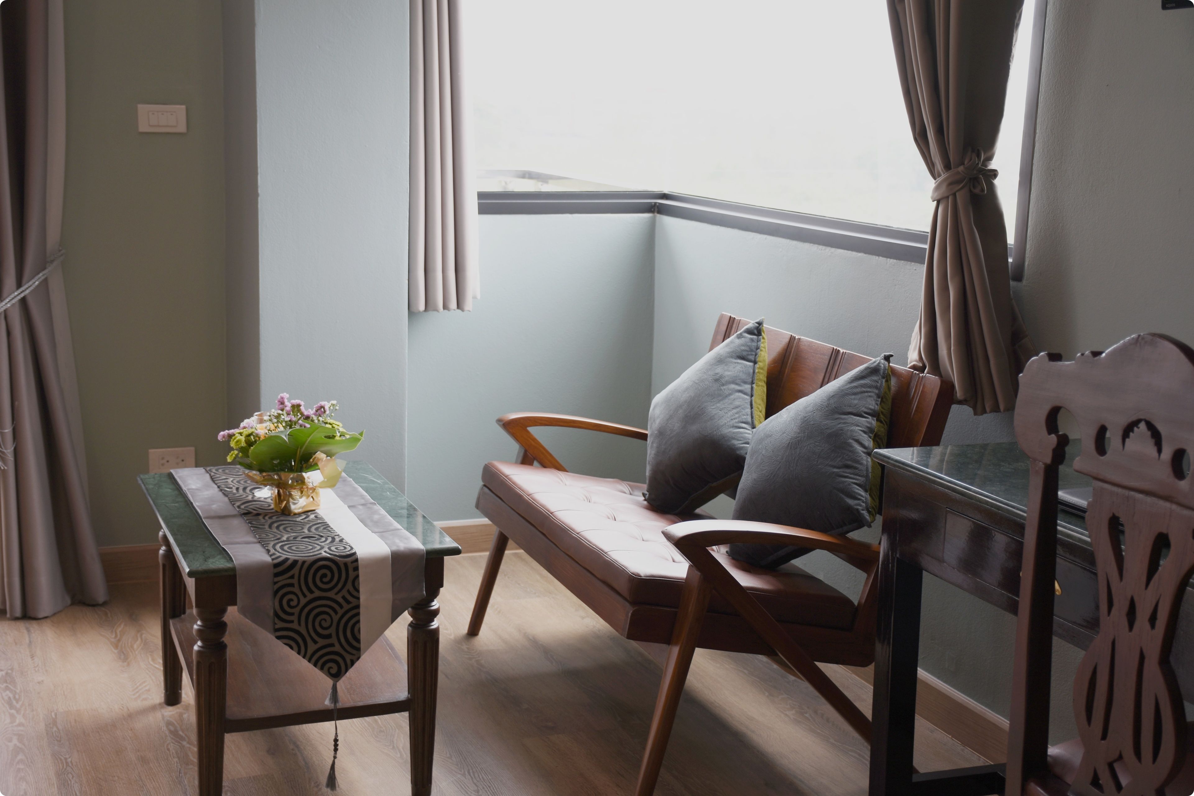 Une pièce d’angle avec un banc en bois, une petite table recouverte d’une nappe à motifs et une fenêtre avec des rideaux.
