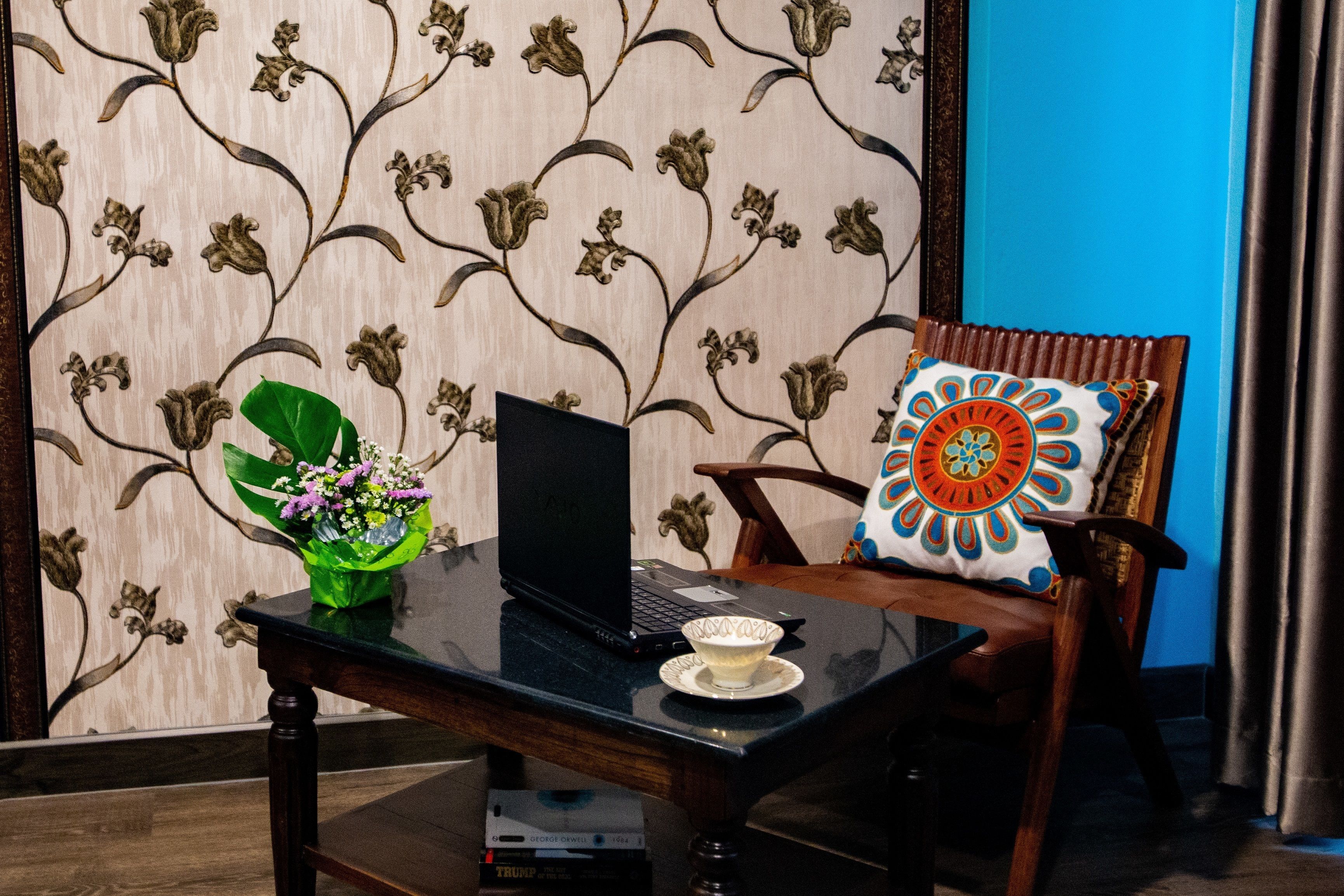 A laptop on a glass table with a decorative bowl, a green plant, and a patterned chair with a colorful pillow.