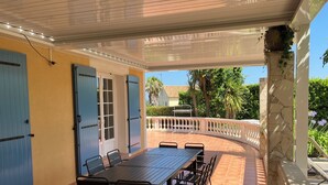 Un patio exterior con mesa y sillas de madera, un área techada y vista a una casa al fondo.