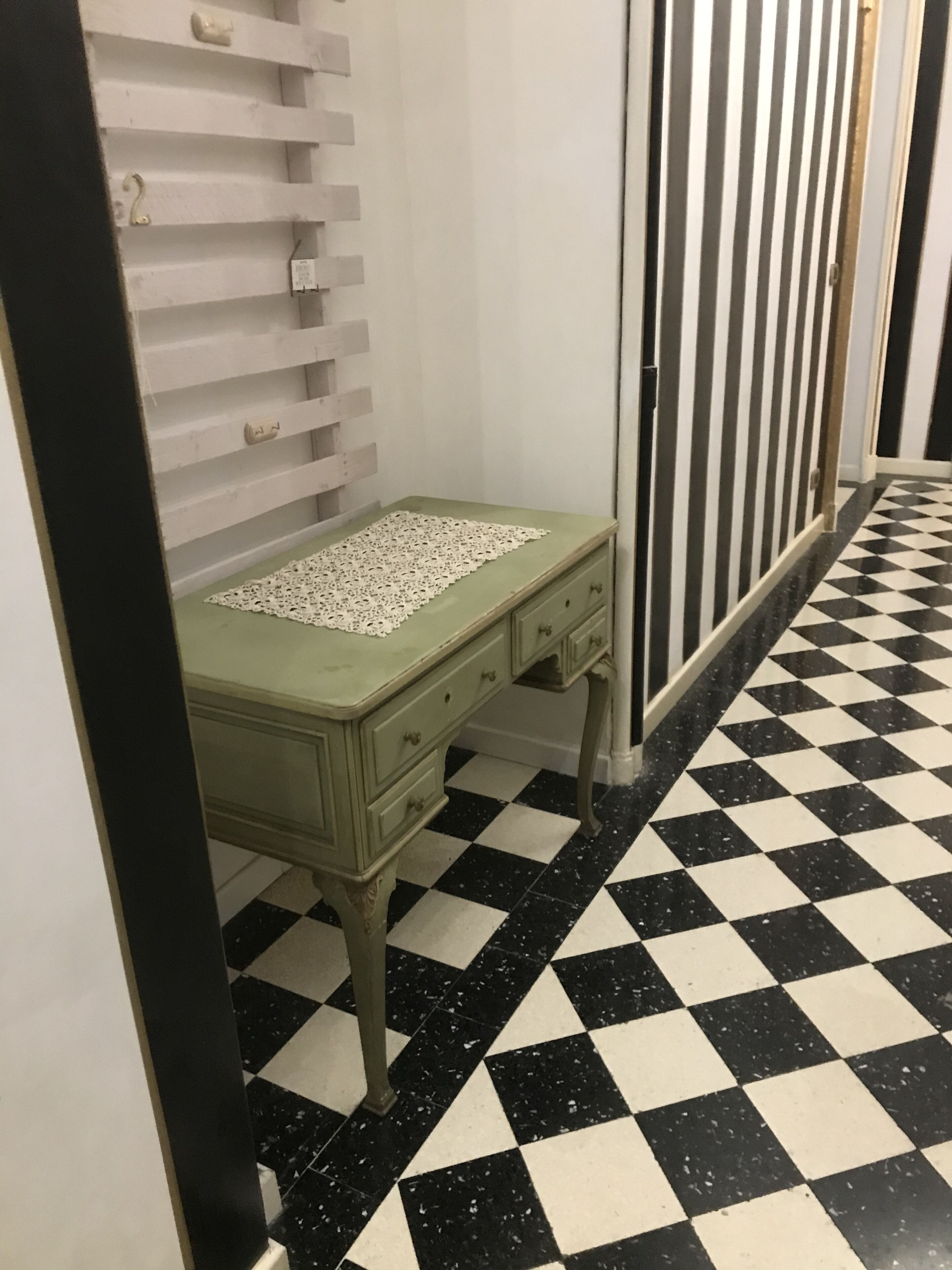 Una stanza con pavimento a scacchi, una cassettiera verde e una tovaglia in pizzo, con una parete bianca e una porta.