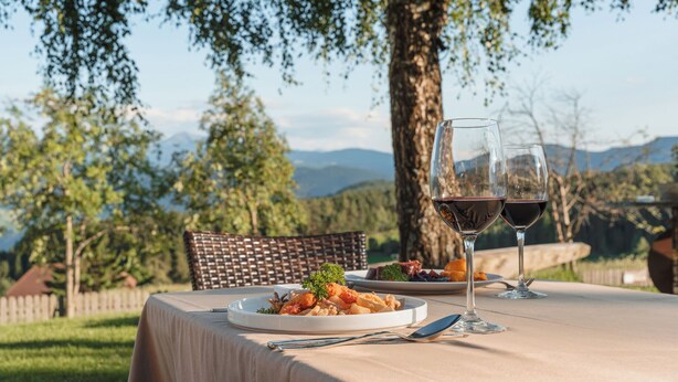 Una mesa de comedor al aire libre con platos de comida y copas de vino tinto, con árboles y montañas de fondo.
