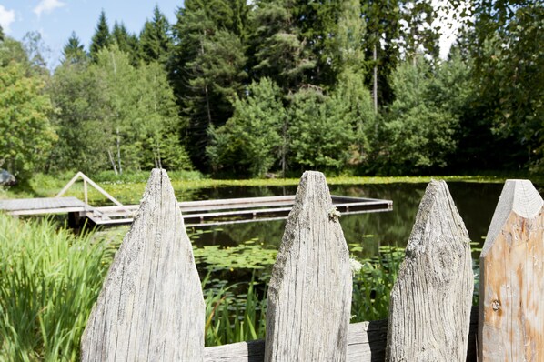 A wooden fence with pointed tops, a wooden dock, and a pond surrounded by dense green trees.