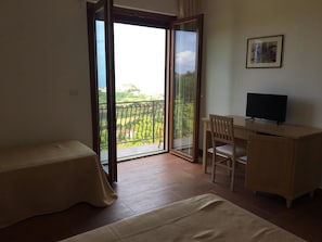 A room with a bed, a desk with a chair, a TV, and a balcony with a view of greenery and buildings.