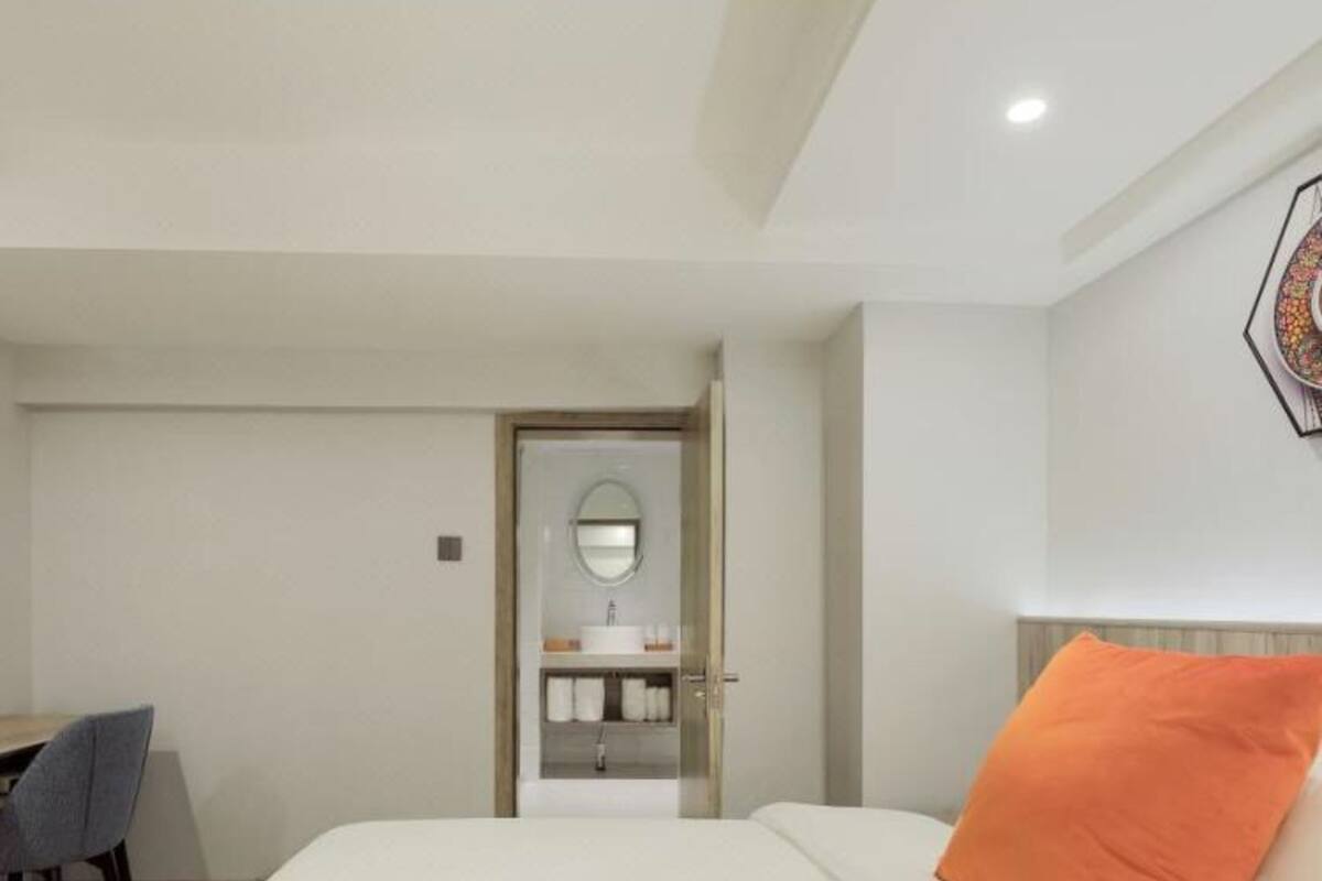 Ein Hotelzimmer mit einem Bett, einem orangefarbenen Kissen, einem Schreibtisch mit Stuhl und einem Badezimmer mit Spiegel und Waschbecken.