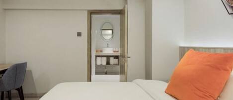 Habitación de hotel con cama, almohada anaranjada, escritorio con silla y baño con espejo y lavamanos.
