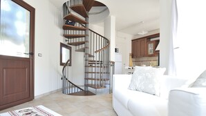 A modern living room with a white sofa, a wooden spiral staircase, and a kitchen in the background. - Residence Bianco Navarrese A (Baunei)
