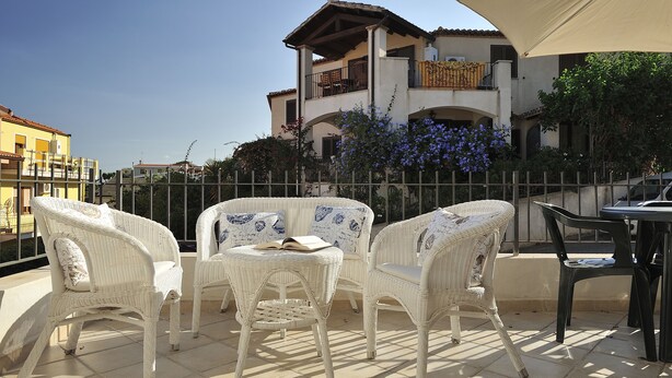 A terrace with wicker furniture and a table, overlooking a building with balconies and a garden. - Residence Bianco Navarrese A (Baunei)
