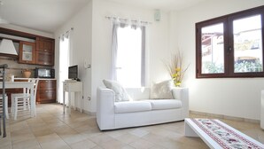 A modern living room with a white sofa, a dining area with a table and chairs, a kitchen with a microwave, and a large window with curtains. - Residence Bianco Navarrese A (Baunei)