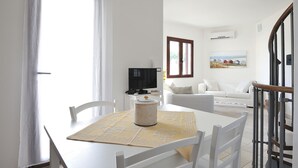 A bright living room with a white sofa, a dining table with chairs, a TV, and a painting on the wall. - Residence Bianco Navarrese A (Baunei)