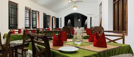 Uma área de refeições com mesas postas, com toalhas de mesa verdes, cadeiras de madeira e guardanapos vermelhos.