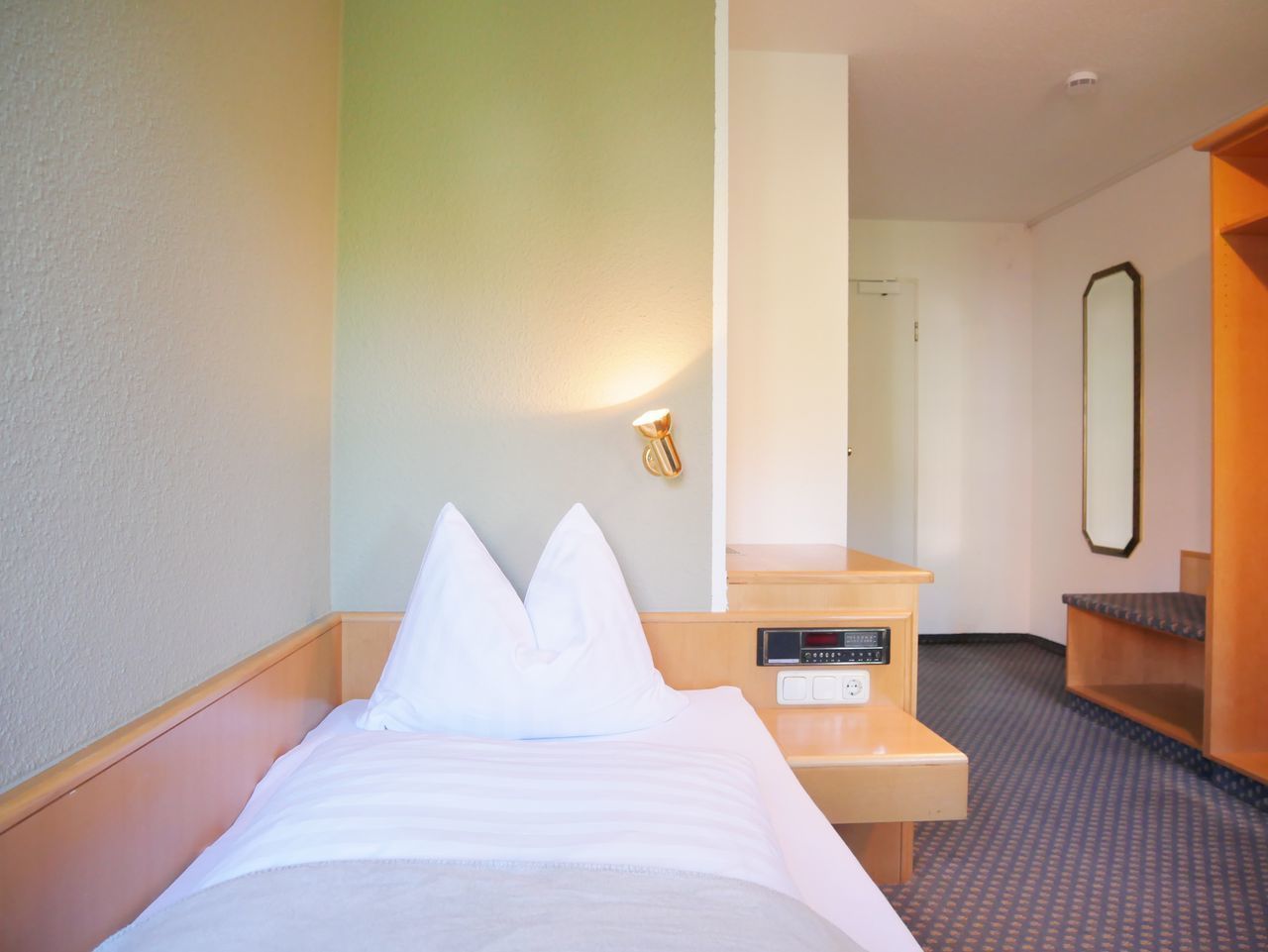 A hotel room with a bed, a bedside table with a control panel, a mirror, and a small seating area.