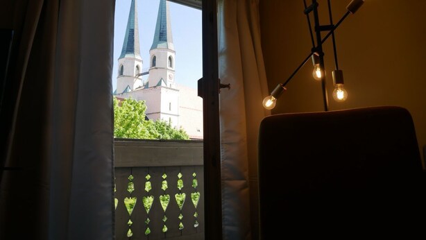 Een kamer met een raam dat uitzicht biedt op een kerk, een stoel en een wandlamp.