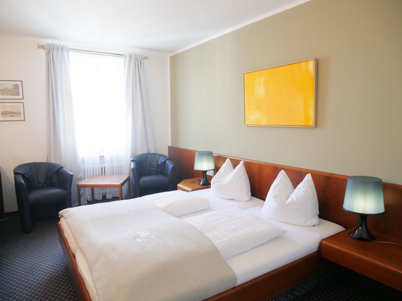 A hotel room with a large bed, two bedside tables, a small table, two chairs, and a window with curtains.