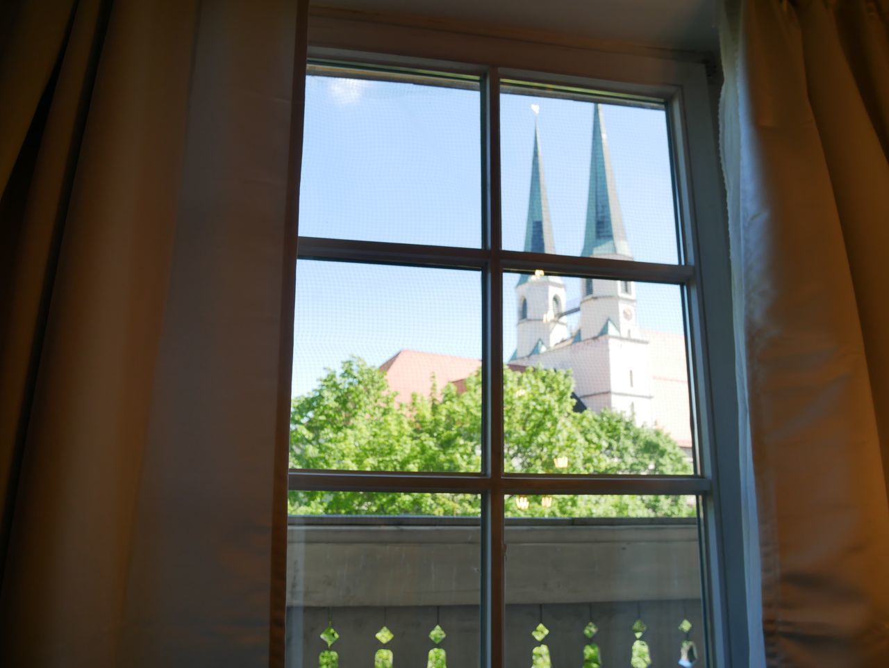 Une fenêtre donnant sur un bâtiment surmonté d’une flèche, avec des rideaux et une clôture ornée de décorations suspendues.