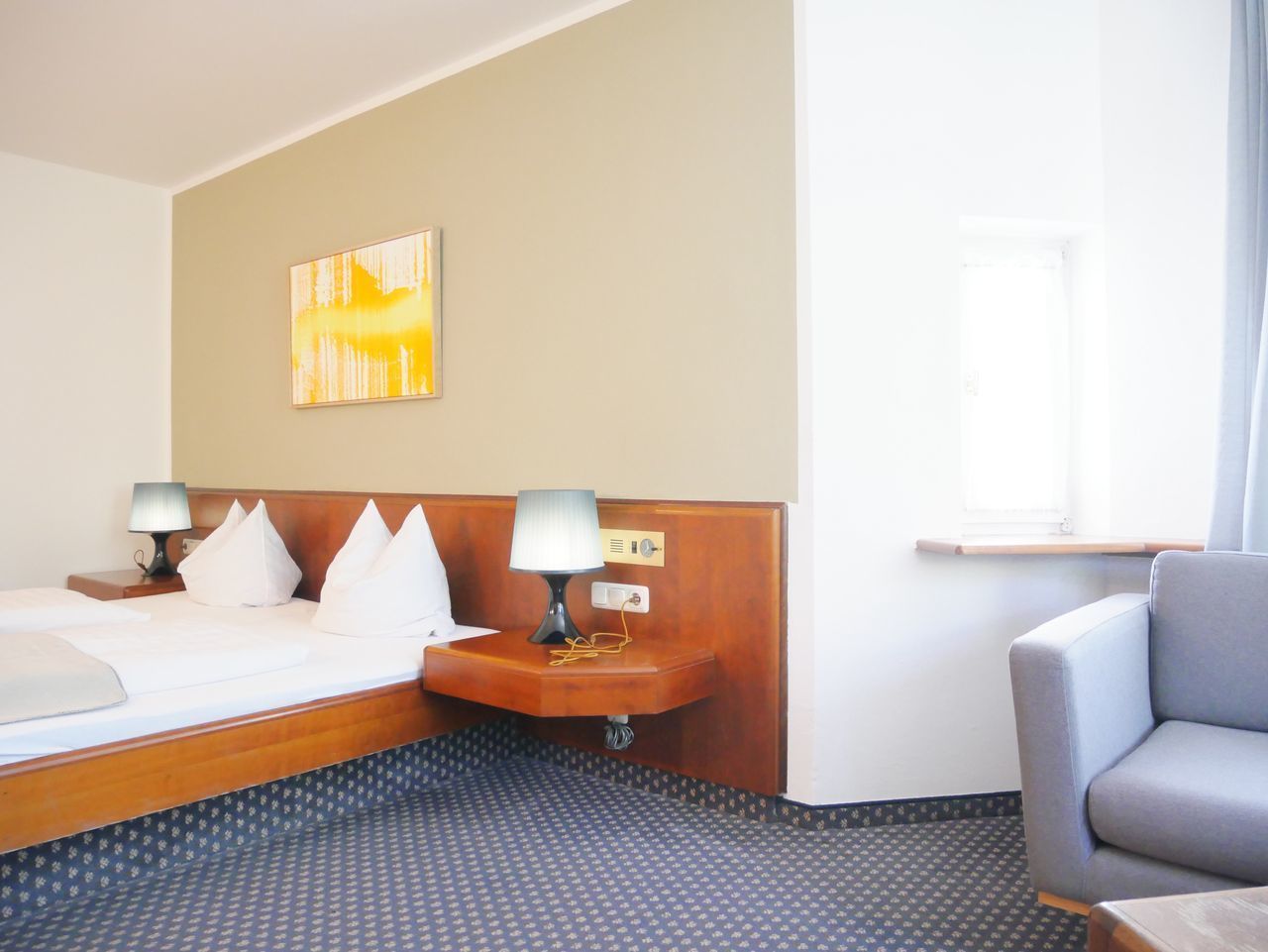 Hotelkamer met een bed, twee nachtkastjes met lampen, een stoel, een raam en een kunstwerk aan de muur.