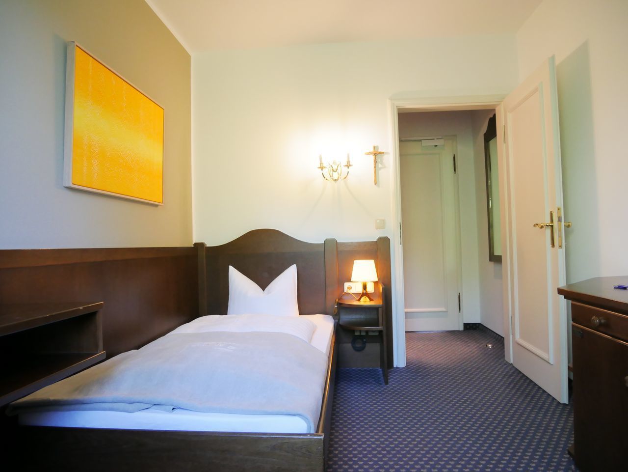 A hotel room with a bed, bedside table, lamp, and a painting on the wall.