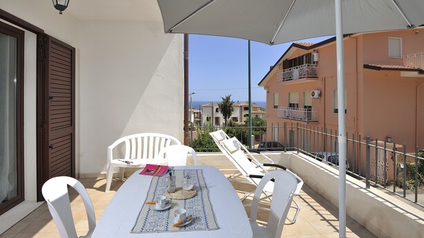 A balcony with a table set for two, white chairs, a white umbrella, and a view of buildings and the sea. - Residence Bianco Navarrese B (Baunei)
