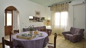 A dining area with a round table covered in a purple tablecloth, surrounded by wooden chairs. There is a window with sheer curtains and a wall-mounted air conditioner. - Casa di Zio Mario A (Baunei)