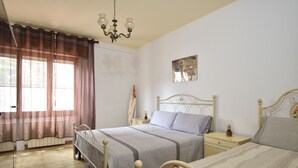 A bedroom with two beds, a chandelier, a bedside table with a lamp, and a window with curtains. - Casa di Zio Mario A (Baunei)