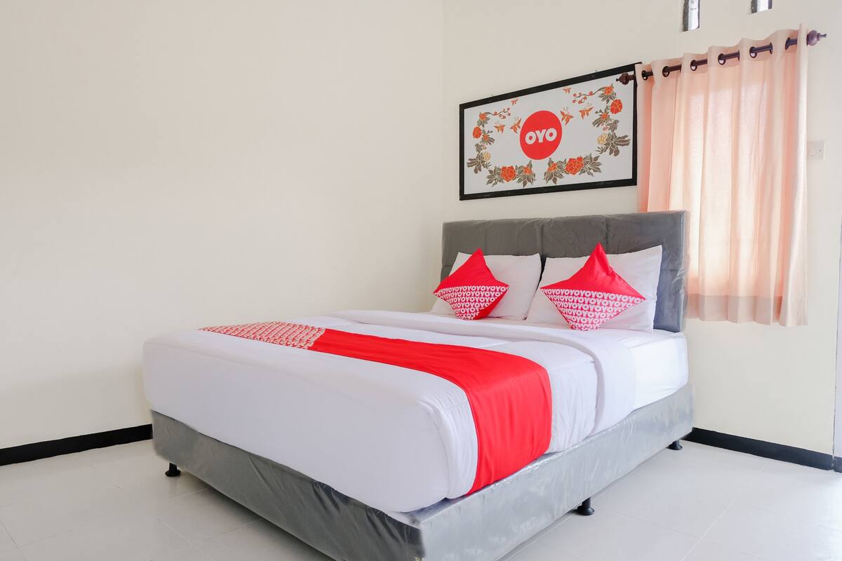 Un lit bien fait, avec une tête de lit grise et une literie blanche, agrémenté de coussins rouges et d’un plaid rouge et blanc. Un tableau floral encadré, sur lequel on peut lire le mot « OYO », est accroché au mur au-dessus du lit.