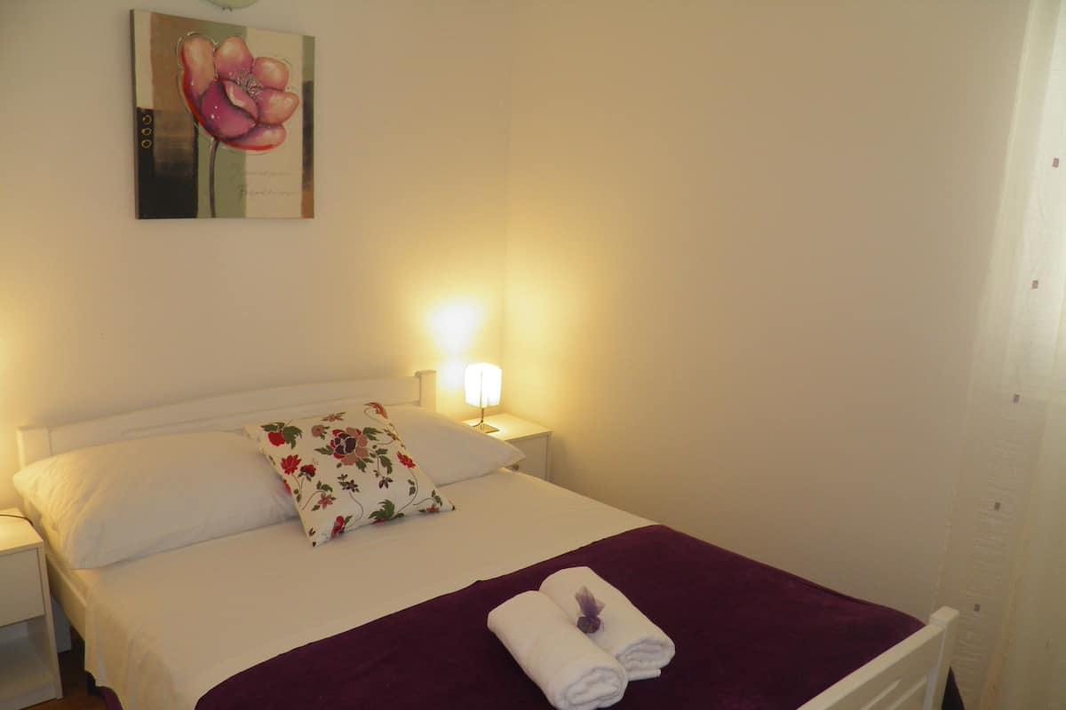 A neatly made bed with a purple blanket, white pillows, and a floral pillow. There is a nightstand with a lamp and a painting of a flower on the wall.