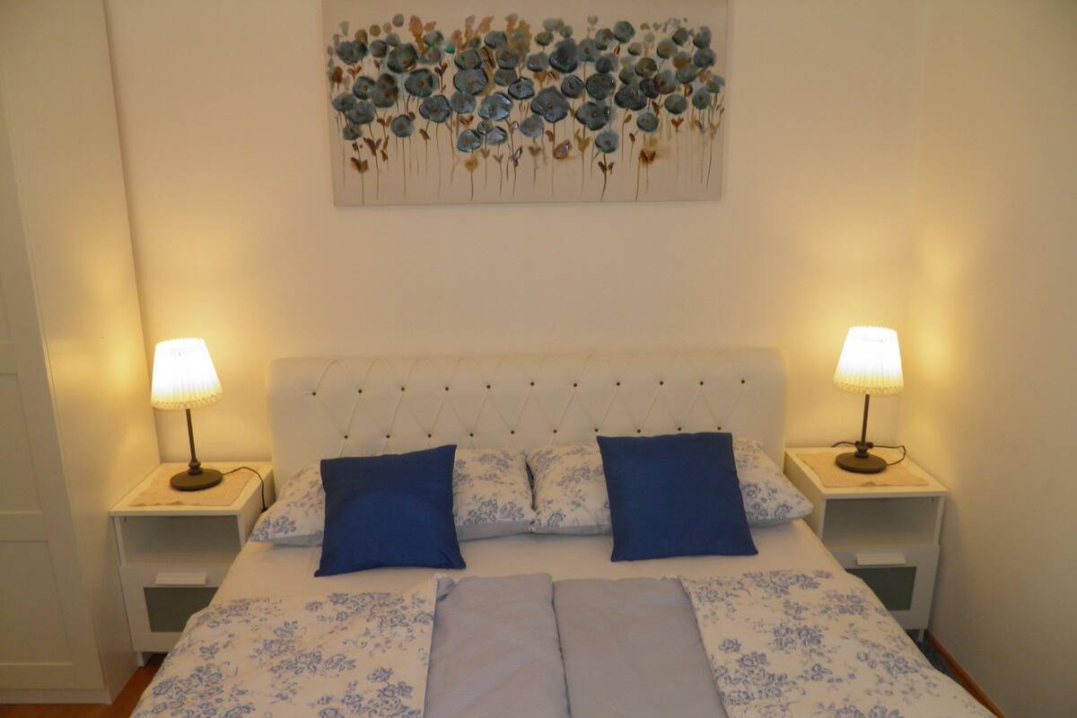 A double bed with a tufted headboard, two bedside tables with lamps, and a floral artwork above the bed.