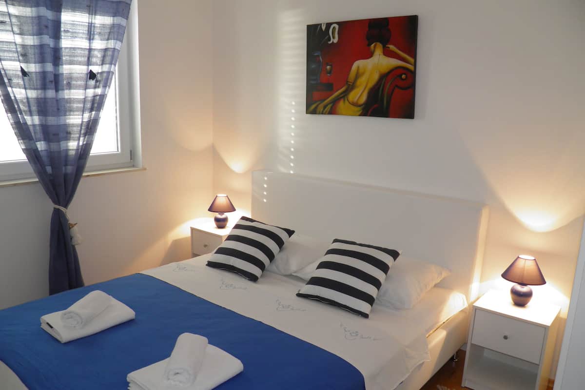 A neatly made bed with a blue bedspread and white pillows, two bedside lamps, and a painting on the wall.
