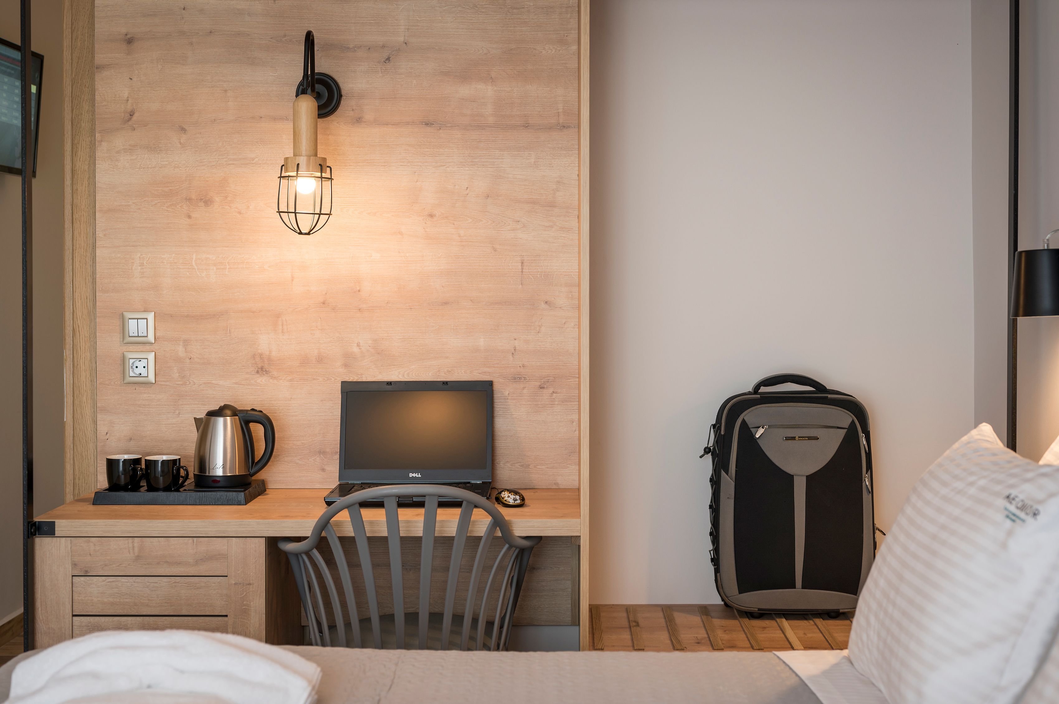 Pieni hotellihuone, jossa on puinen työpöytä, kannettava tietokone, vedenkeitin ja matkalaukku.