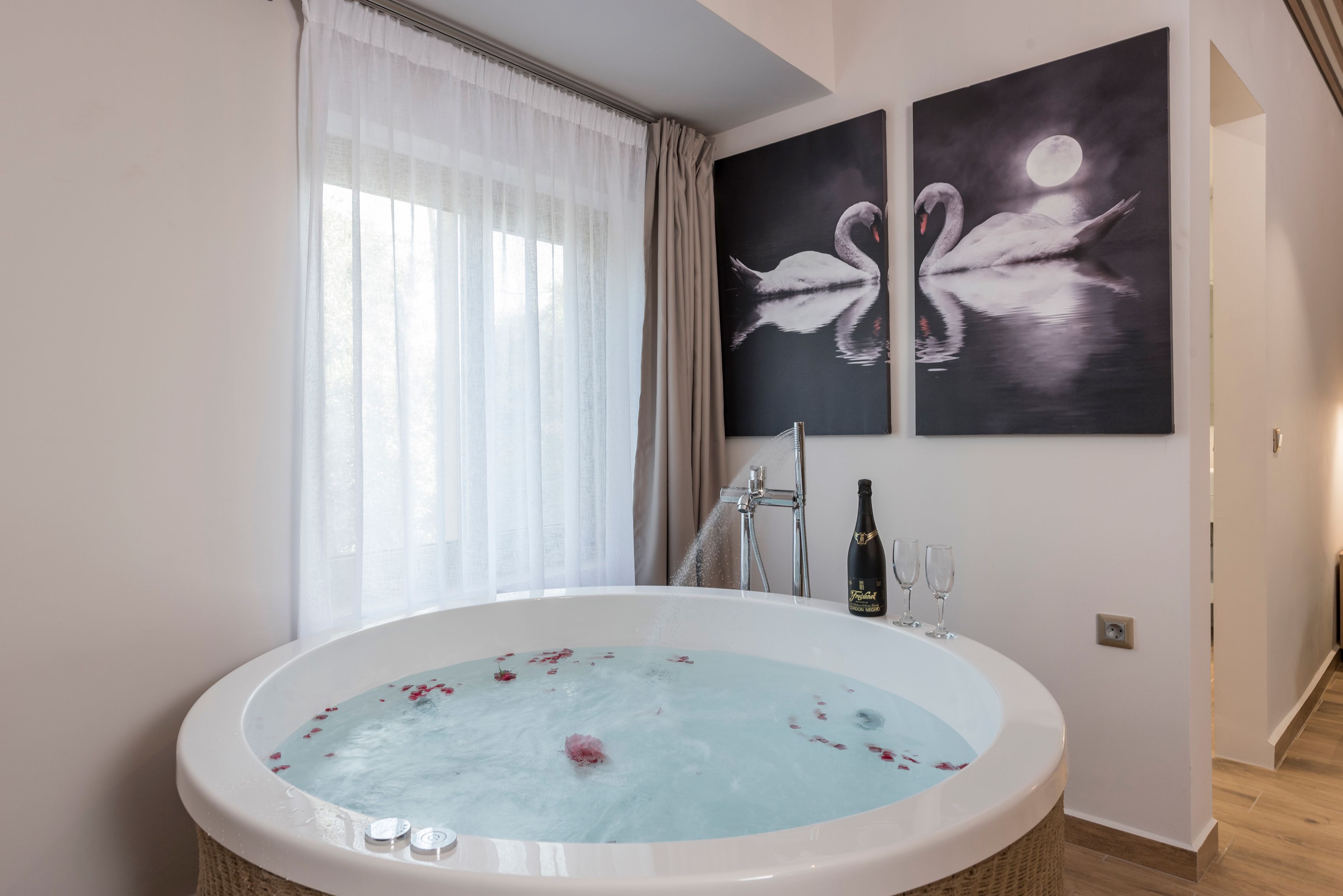 Une salle de bain moderne avec une baignoire îlot remplie d’eau et de pétales de rose, une grande fenêtre ornée de rideaux légers et deux tableaux représentant des cygnes, accrochés au mur.