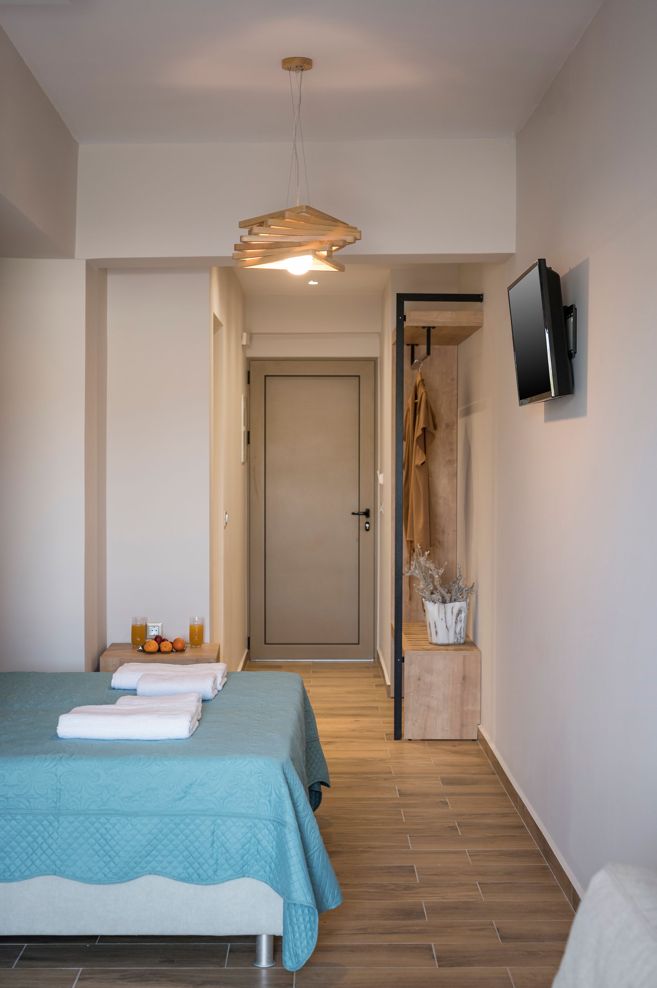 A modern hotel room with a bed, a flat-screen TV, a wooden floor, and a hallway leading to another room.