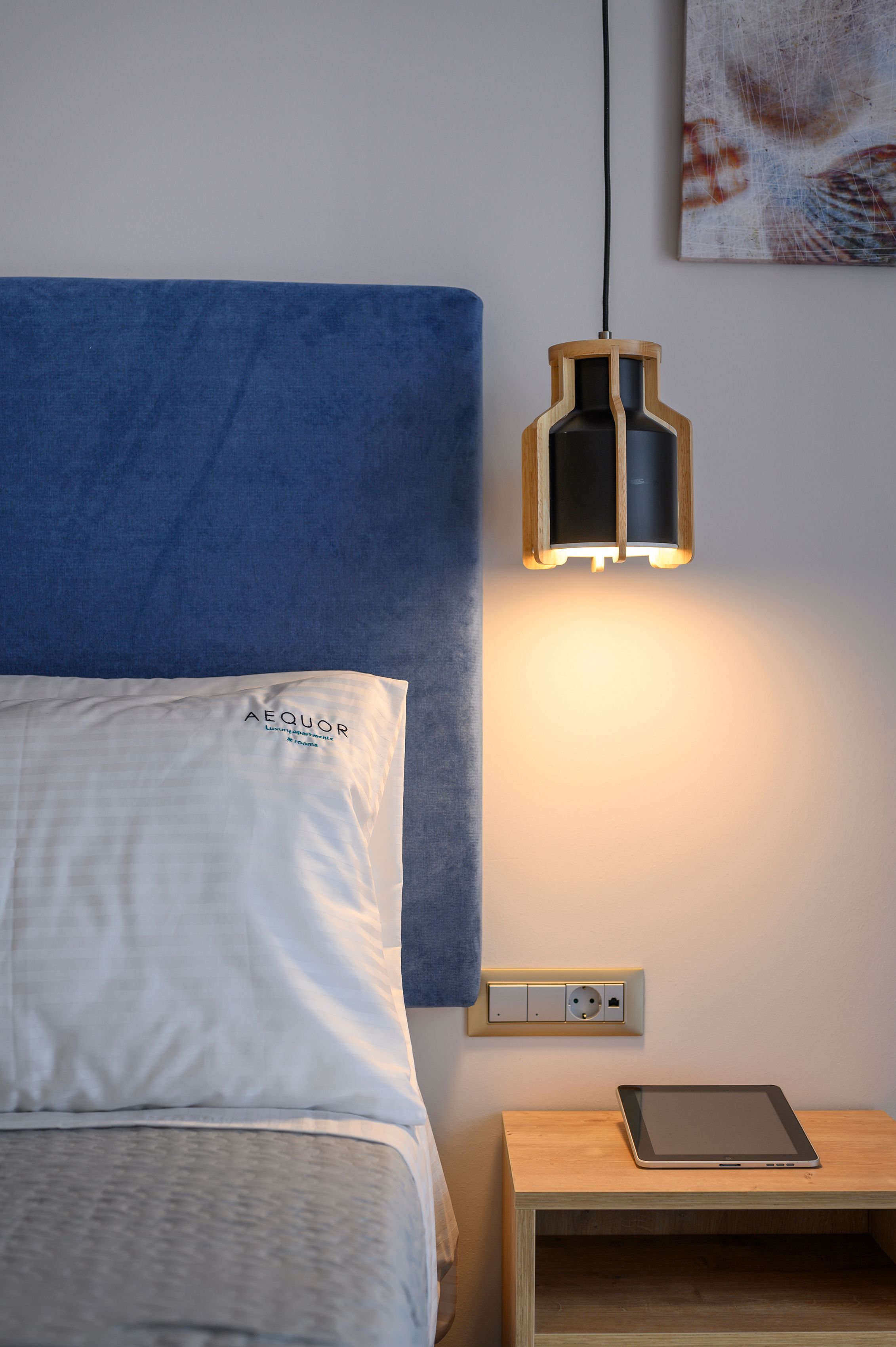 A neatly made bed with a blue headboard, a bedside table with a tablet, and a wall-mounted light fixture.