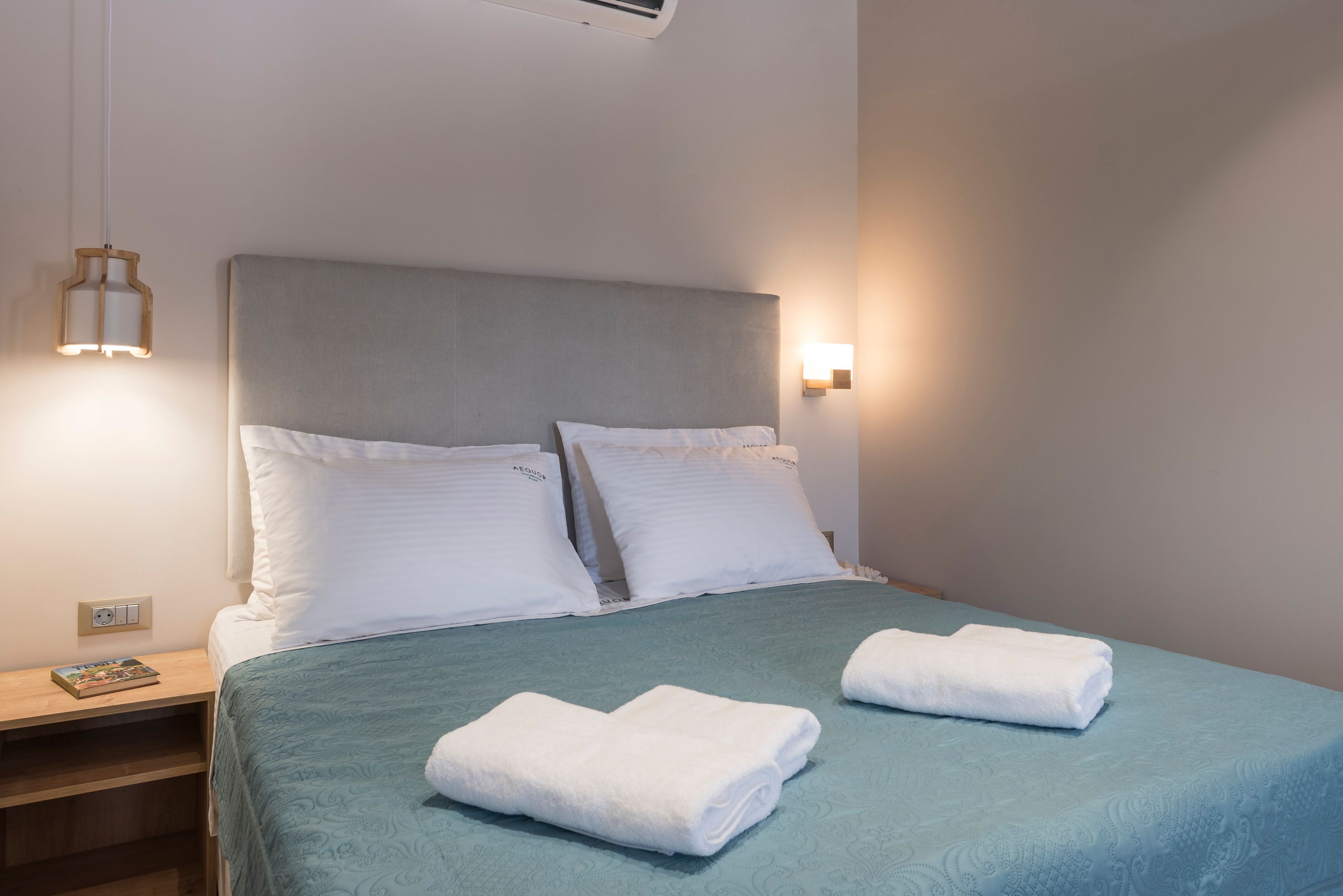 A neatly made bed with white linens and two folded towels, a bedside table with a book, and a wall-mounted light fixture.