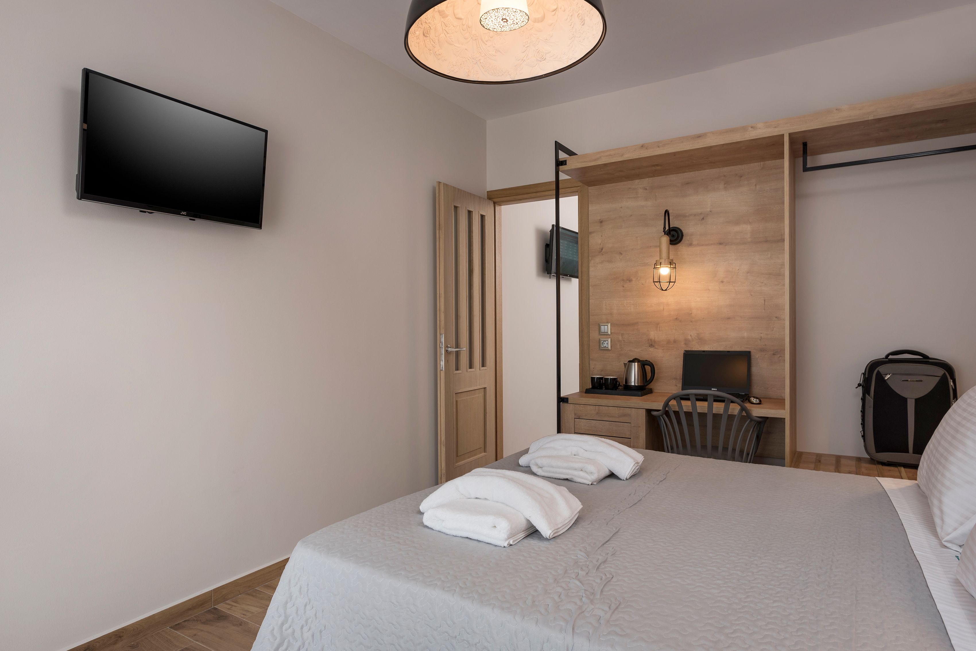 A hotel room with a bed, a flat-screen TV mounted on the wall, a desk with a computer, and a kitchenette area.