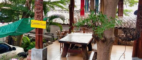 A covered outdoor seating area with a table and chairs, a sign indicating a common room, and various potted plants.