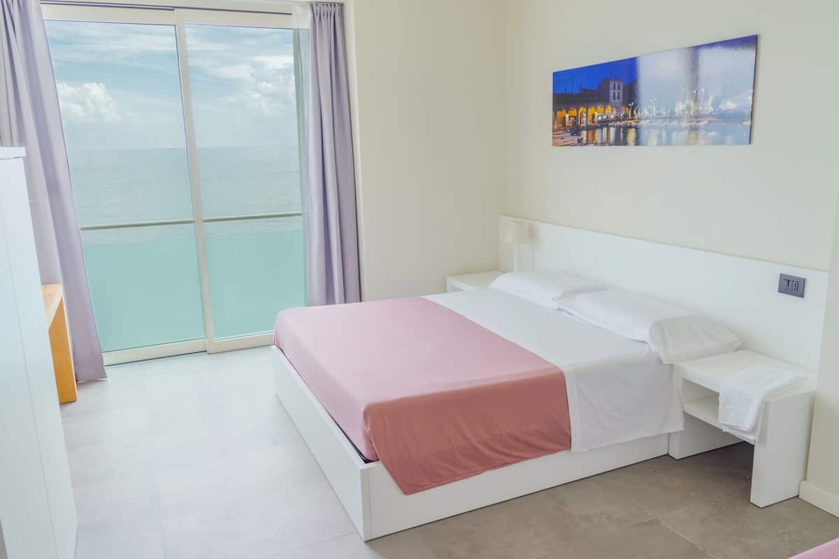 A hotel room with a bed, a nightstand, a painting, and a view of the sea through a large window.