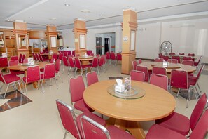 A spacious dining area with multiple round tables and red chairs, featuring a central column with a fan and a glass-top table. - Halim Hotel (Tanjung Pinang)