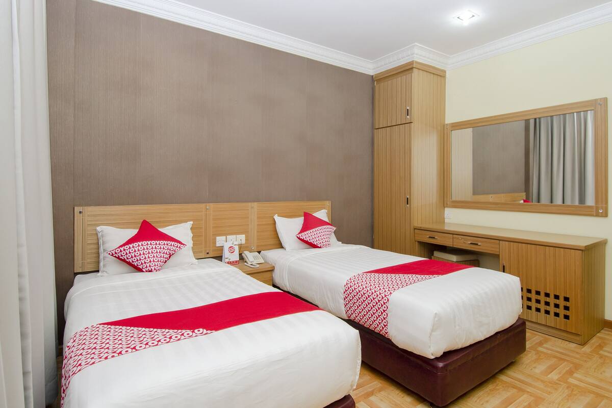 A hotel room with two beds, each with white bedding and red accents, a wooden headboard, a desk with a mirror, and a wardrobe.