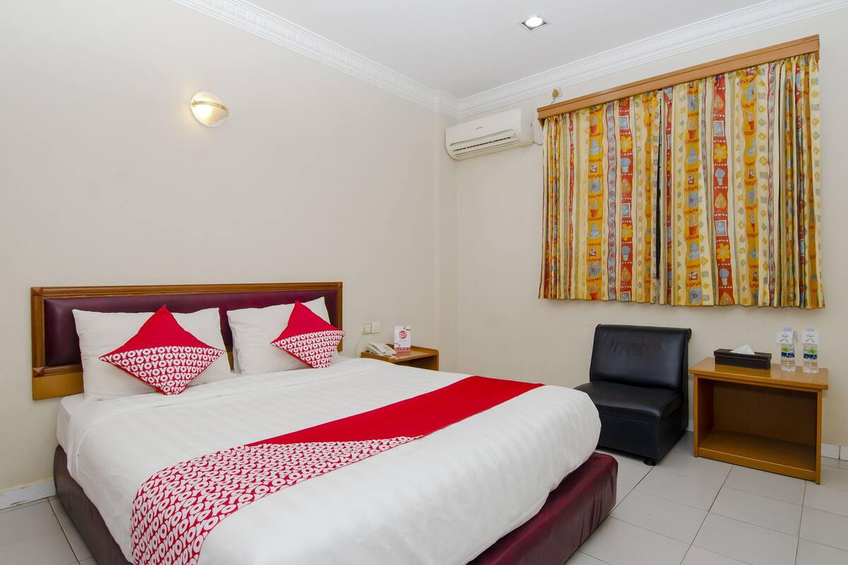 A hotel room with a large bed, a bedside table, a chair, an air conditioning unit, and a curtain with a colorful pattern.