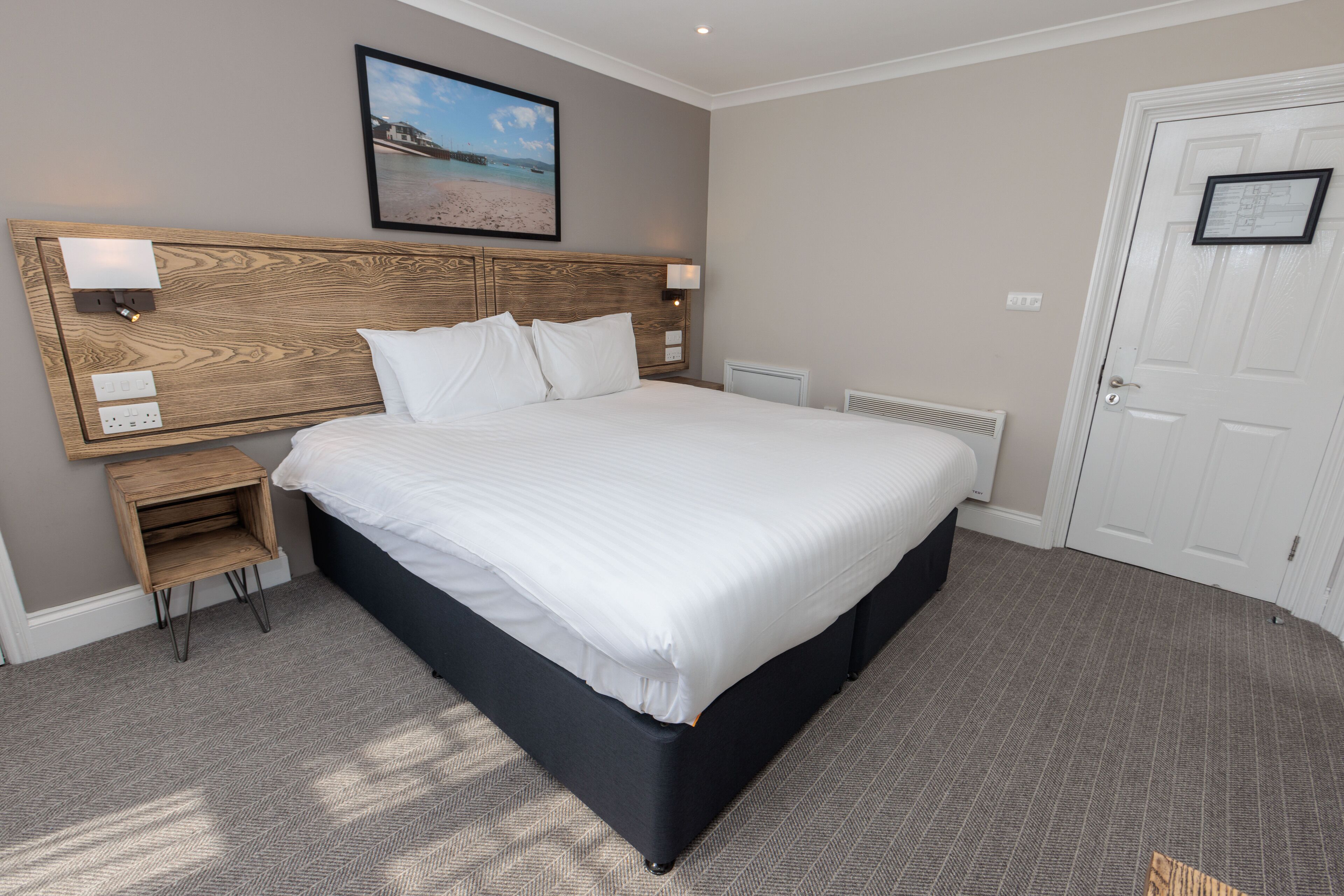 Quarto de hotel com uma cama grande, cabeceira de madeira, mesa de cabeceira e um quadro na parede.