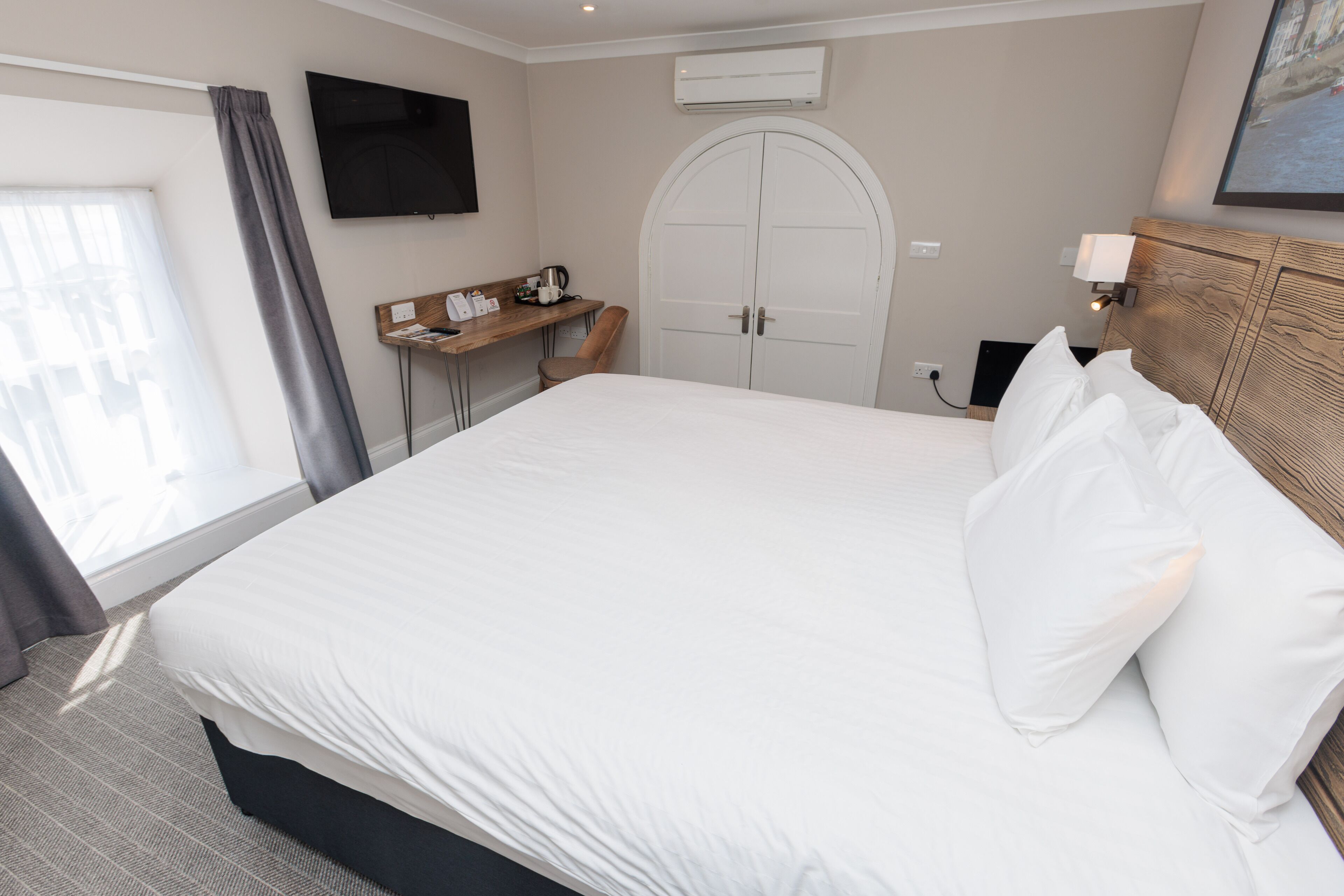 A hotel room with a large bed, a desk with a chair, a TV mounted on the wall, and a door with a coat rack.