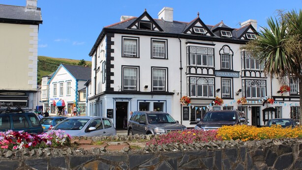 A traditional building with a sign 'DOVEY INN' indicating a hotel. There are parked cars in front, and a stone wall with flowers in the foreground. - Dovey Inn (Aberdovey)