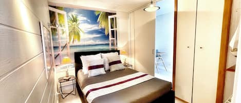 A bedroom with a bed, a nightstand, a lamp, a wardrobe, and a view of the ocean through a window.