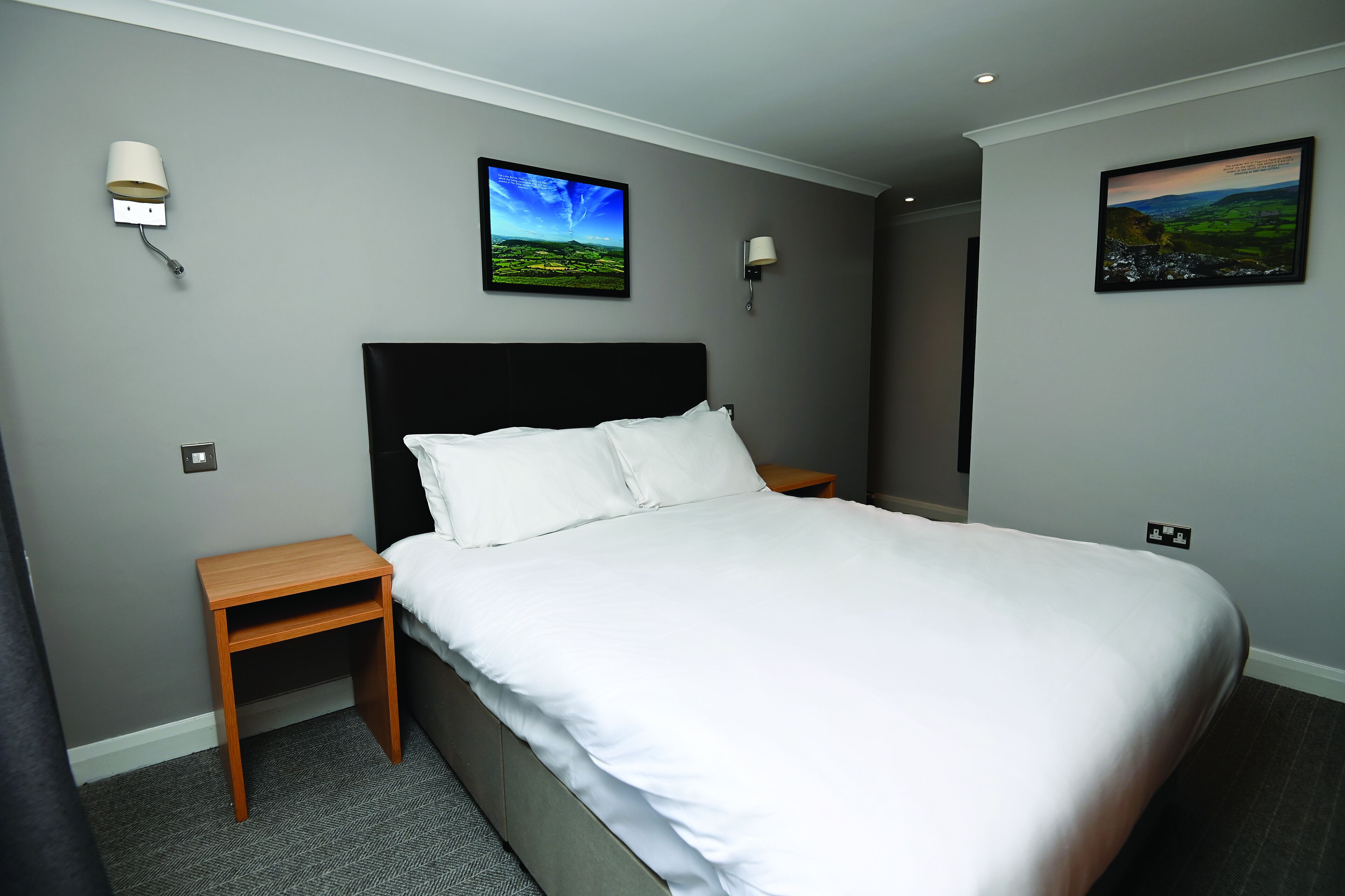 Una cama bien hecha en una habitación de hotel, con una pared gris, dos lámparas fijadas a la pared y dos cuadros enmarcados.