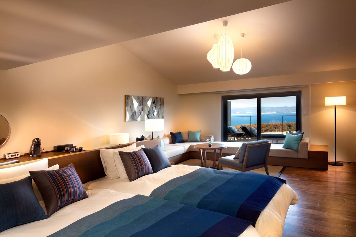 A modern hotel room with a large bed, a sofa, a small table, and a view of the outdoors.